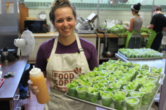 Meghan, wearing a FoodCorps tee and apron, holds a large tray filled with plastic cups of salad