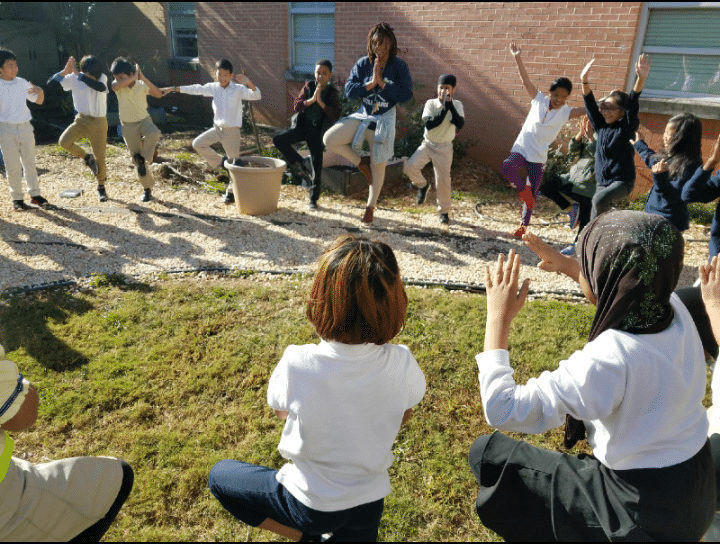 FoodCorps alum Zashe Cockett-Demings (GA '18) stands in a circle of students outdoors; some are balancing on one foot, others are holding their hands out. Movement in lessons encourages positive mental health among students.