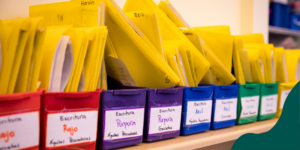 A row of colorful boxes full of papers, labeled with colors and terminology in Spanish, at a school where many students are Latine.