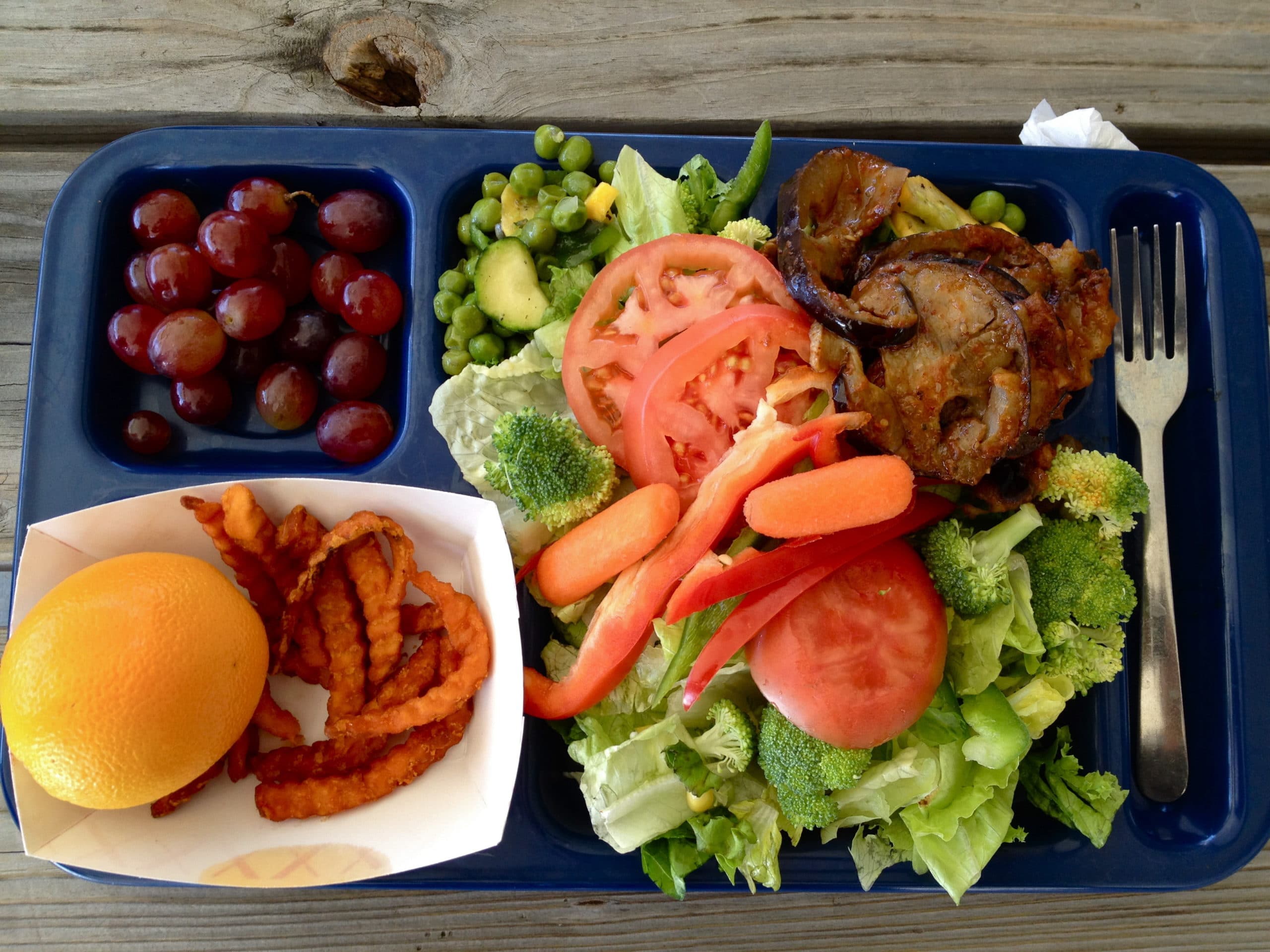 A lunch tray in Arkansas featuring an array of school food items, including fresh broccoli, purple grapes, sweet potato fries, and an orange.