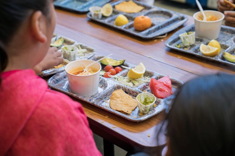 A distant view of school lunch trays on a table, with children sitting at the table and facing away. The NSLP helps make sure all kids can be fed at school.