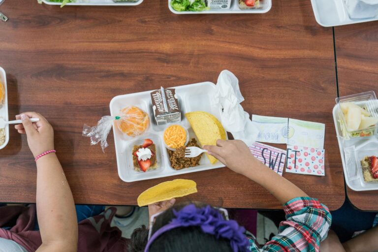 An overhead photo of a child with dark hair and a plaid shirt sitting at a lunch table and scooping meat into a taco shell.