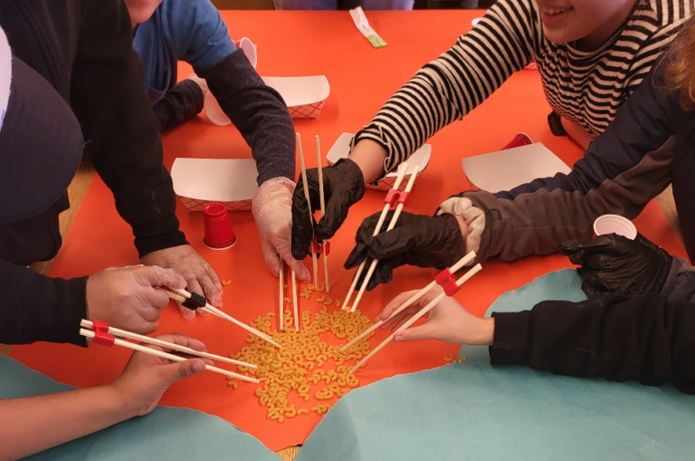 A close-up of students’ hands, some wearing food prep gloves and some not, practicing using chopsticks by picking up dry pieces of macaroni.