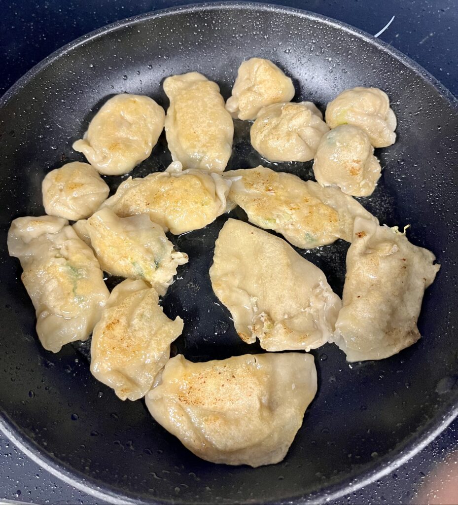 Close-up of more than a dozen golden-brown dumplings frying in an oiled pan. 