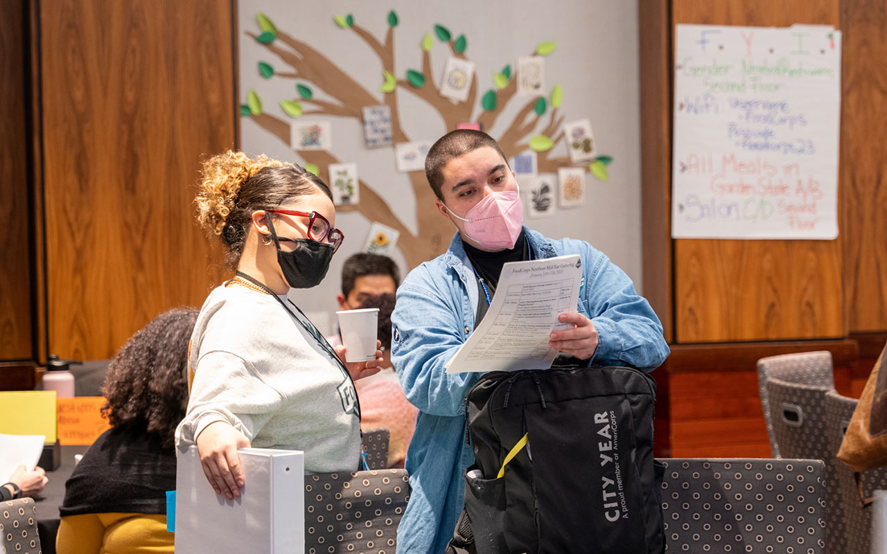 Service members Shay Brooks and Isa Atencio, both wearing FoodCorps gear, stand at a round table, both looking intently at a paper. On the table in front of them are markers, papers and binders; in the background are other service members, as well as flip chart paper displayed on the wall.