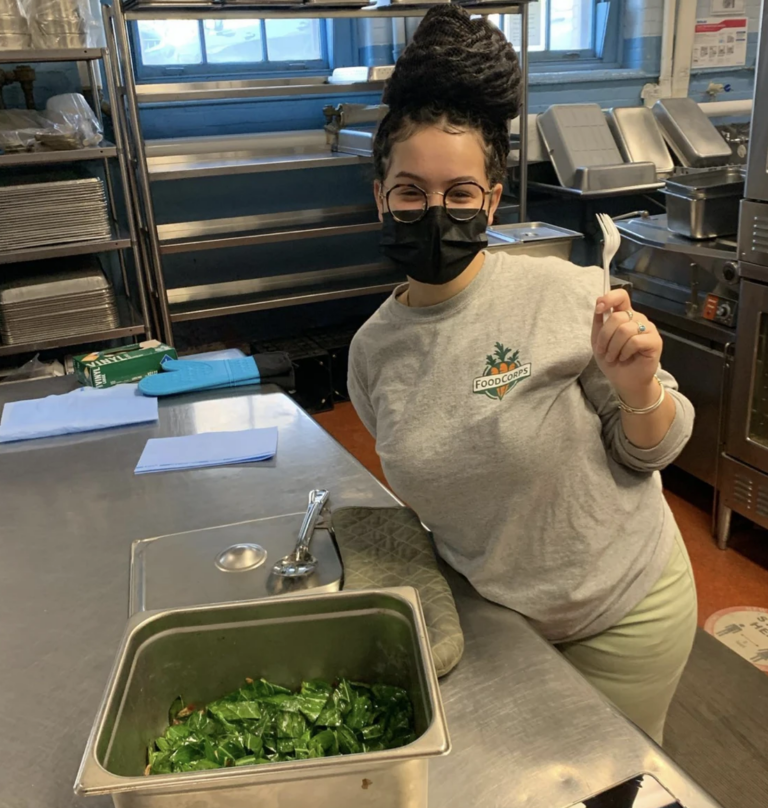 Shay Brooks, wearing a grey FoodCorps sweatshirt, holds up a plastic fork while standing in the cafeteria in front of a container of collard greens.