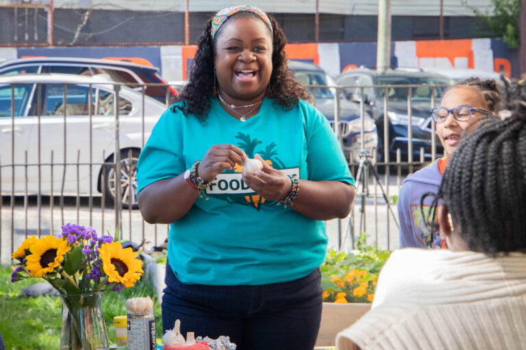 Bridgette Byrd, a Black woman with dark curly hair pulled back with a headband and wearing a green FoodCorps shirt and jeans, holds a garlic bulb while smiling and talking to a student whose back is to the camera. A student wearing purple glasses stands next to Bridgette, and around them are grass, flowers, and other garden materials.