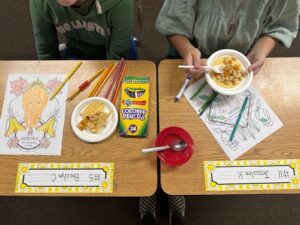 An overhead image of two students’ desks side by side. On each desk is a small taco on a plate and a partially-colored art page with colored pencils scattered nearby. One student is wearing a green hoodie and the other is wearing a flowy green shirt; neither of their faces are visible. These lessons help contribute to creating a sense of belonging for all students.