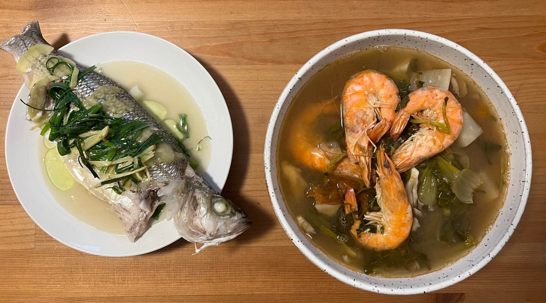 Two dishes of food, side by side. The plate on the left has a steamed fish with wilted scallions on top. The bowl on the right has soup with shrimp and vegetables.