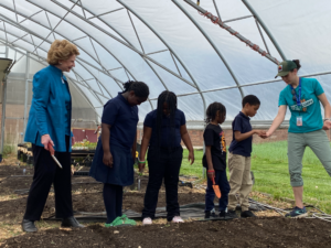 Michigan Sen. Debbie Stabenow stands next to four students and a FoodCorps service member in a greenhouse. Stabenow and the students are holding trowels, preparing to plant.