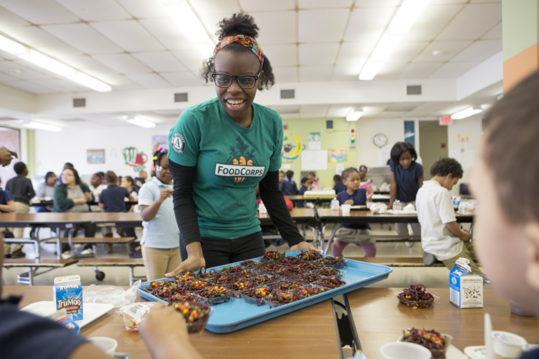 A smiling FoodCorps service member, with dark skin and dark curly hair tied back in a headband, holds a blue tray of snack cups in front of a table of students.