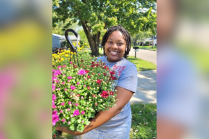Alumni farmer LaBria Lane is standing outside and smiling, wearing a grey t-shirt and holding a big planter of colorful flowers.