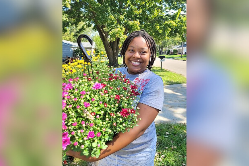 Alumni farmer LaBria Lane is standing outside and smiling, wearing a grey t-shirt and holding a big planter of colorful flowers.