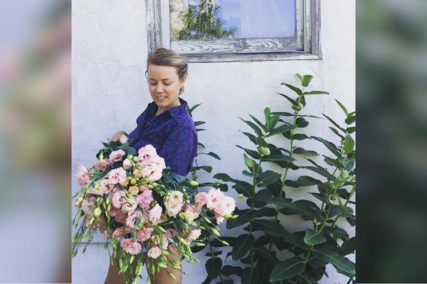 Alumni farmer Molly Schintler stands against a white wall and smiles, looking down at a large bouquet of flowers.