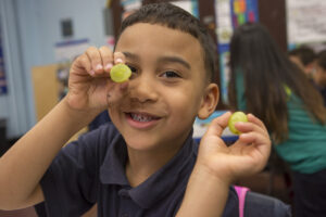 A student wearing a navy blue polo shirt holds up two green grapes and smiles for the camera. The Fresh Fruit and Vegetable Program makes fruit and veggie snacks more accessible during the school day.
