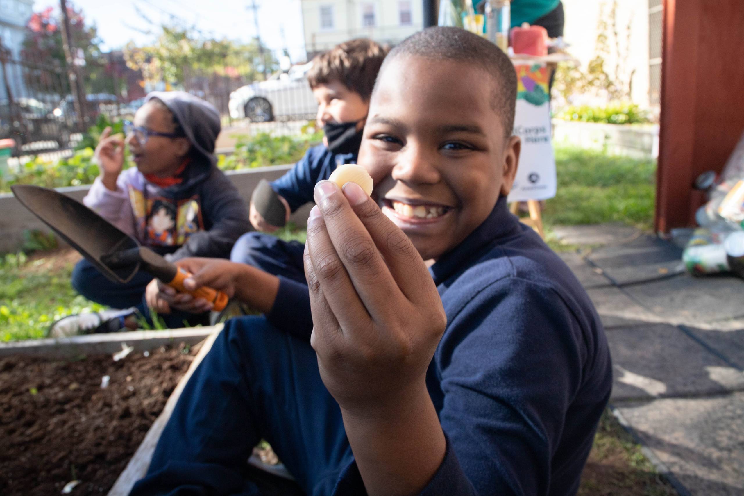 A smiling student wearing a navy blue shirt holds up a freshly-harvested garlic clove to the camera.