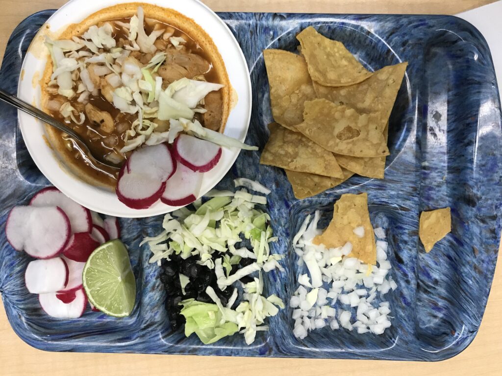 A blue lunch tray with a bowl of pozole and sides of tortilla chips, diced onions, sliced radishes, shredded lettuce, and black beans
