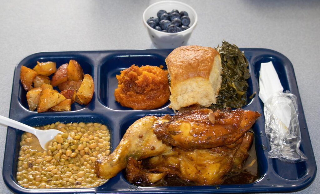 A blue lunch tray holding baked chicken, lentils, sweet potato mash, greens, roasted potatoes, and a roll, with a small cup of blueberries on the table nearby