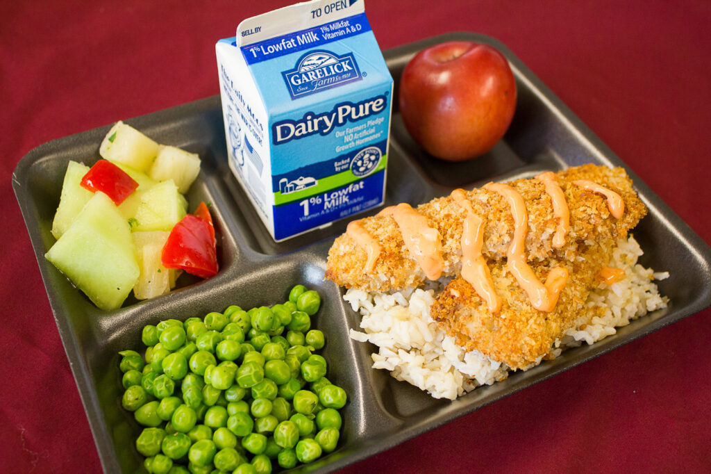 A black lunch tray holding coconut crusted fish over rice, peas, chopped veggies, an apple and a milk carton