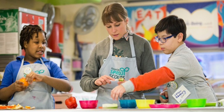 A FoodCorps member stands at a table with two students during a cooking class, measuring out ingredients. Involving kids in cooking fall recipes can help build their curiosity and interest in nourishing food.