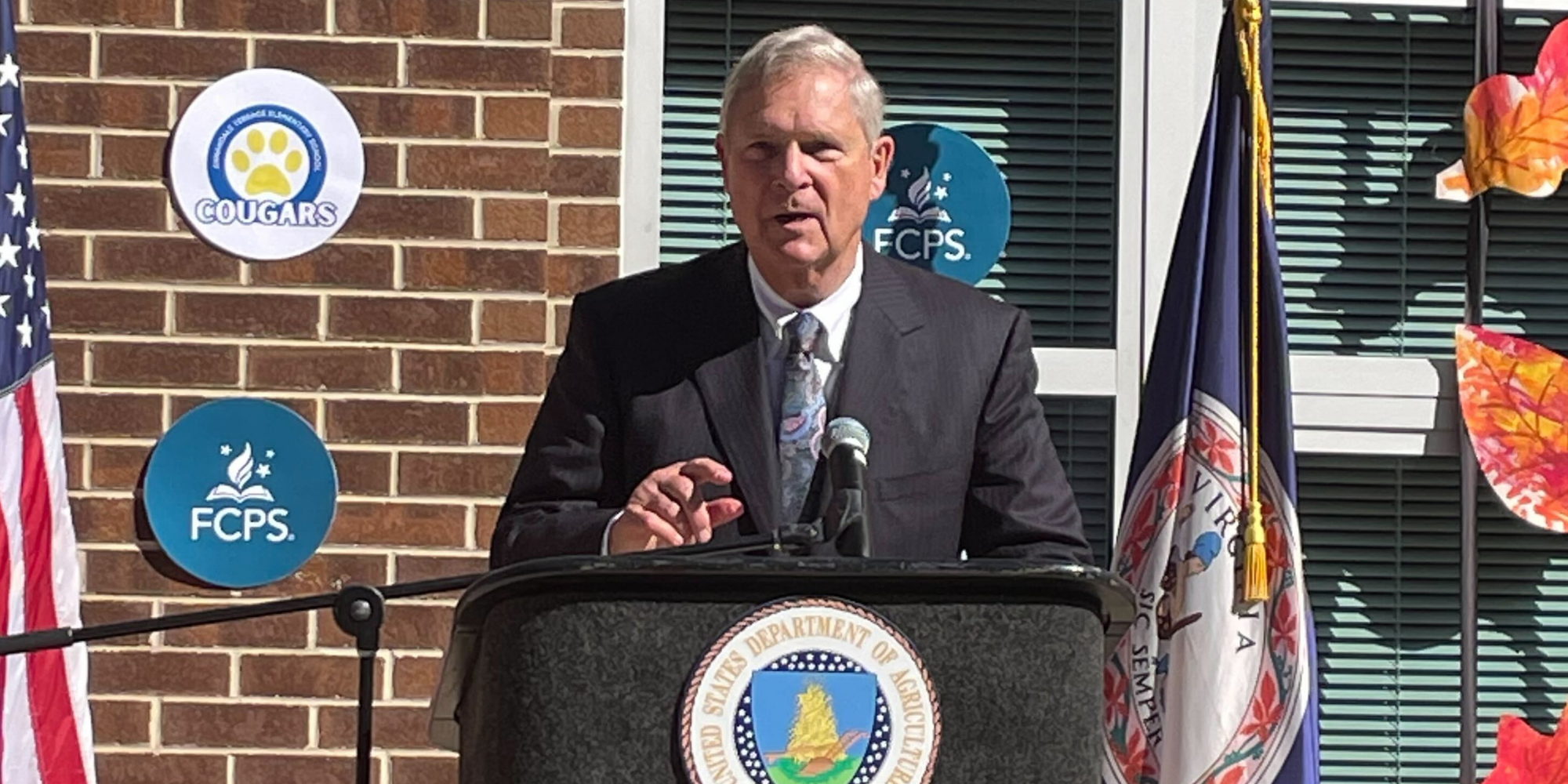 USDA Secretary Tom Vilsack, wearing a black suit and standing at a podium in front of a brick building, announces new grant opportunities during National School Lunch Week at Annandale Terrace Elementary School in Virginia, an important moment for school food policy.