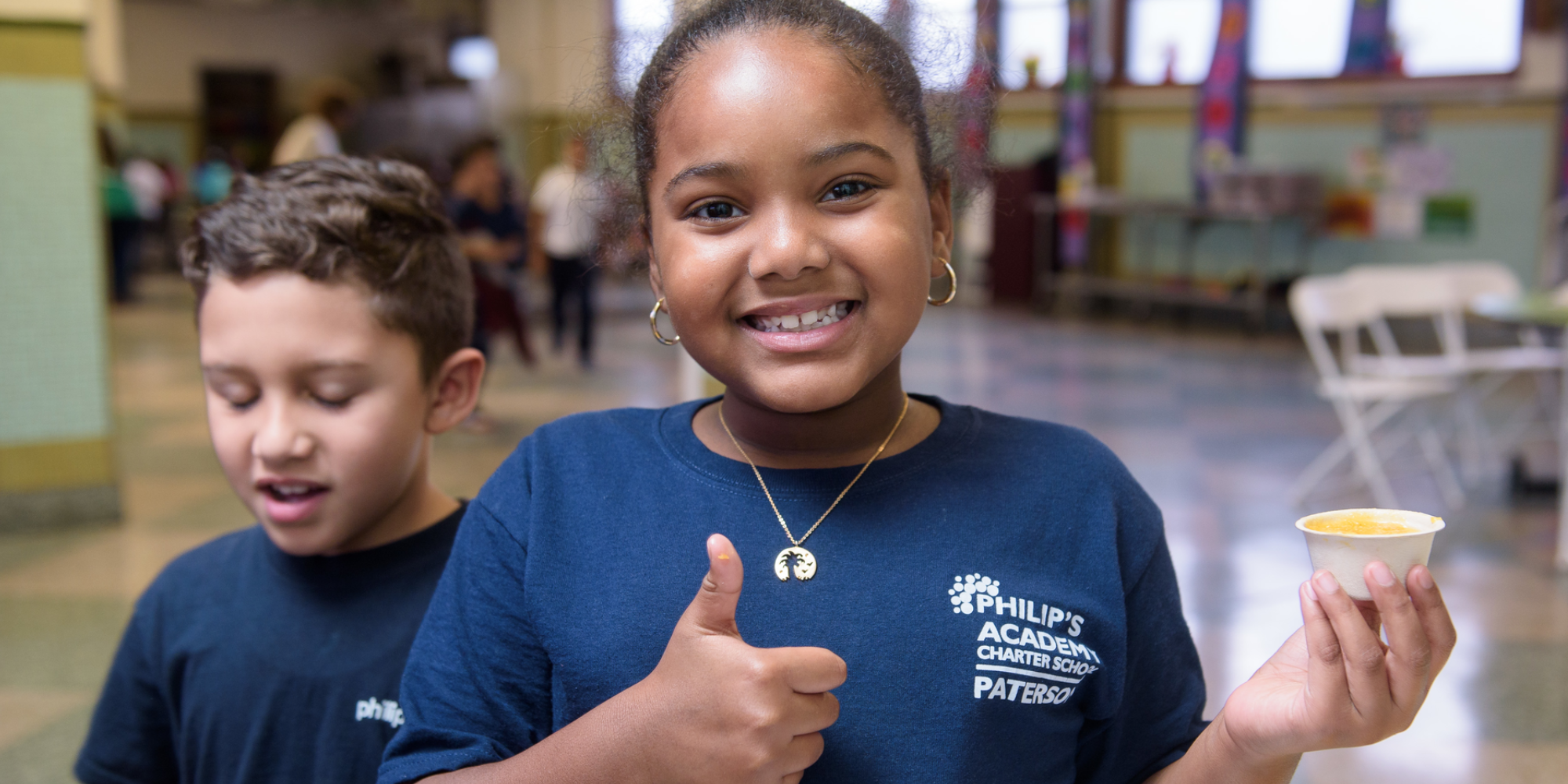A smiling elementary-aged student wearing a blue school t-shirt holds a sample cup of butternut squash soup and gives a thumbs-up. A student next to them reacts to their own sample of squash during the taste test.