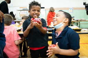 In a Michigan lunchroom, two elementary-age students wearing navy blue polo shirts with short dark hair stand smiling and interacting with each other. Each is holding a red apple, and one is wearing a mask below their chin. Students in the background are seen eating their lunch and enjoying each other’s company. As we share in our policy year in review, Michigan is one of several states that has adopted free school meals in the 2023-2024 school year.