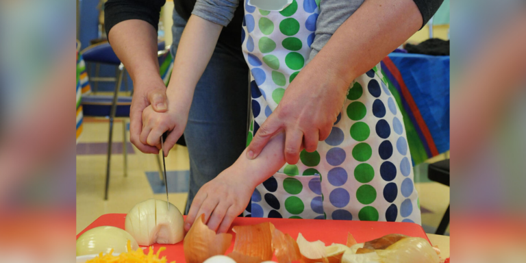 An adult guides a child in chopping an onion. The camera is focused on their hands and arms; their faces are not visible. Families can incorporate healthy habits by cooking and choosing foods together.