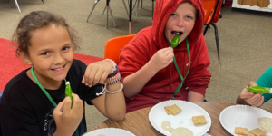 Two students sit at a round table while sampling fresh spring recipes: crackers, green peppers from the garden, and yogurt dip on paper plates in front of them. The student on the left is wearing a black t-shirt and several bracelets and holding up a pepper, smiling; the student on the right is wearing a red hoodie with the hood up and putting a pepper in their mouth with a goofy look on their face.