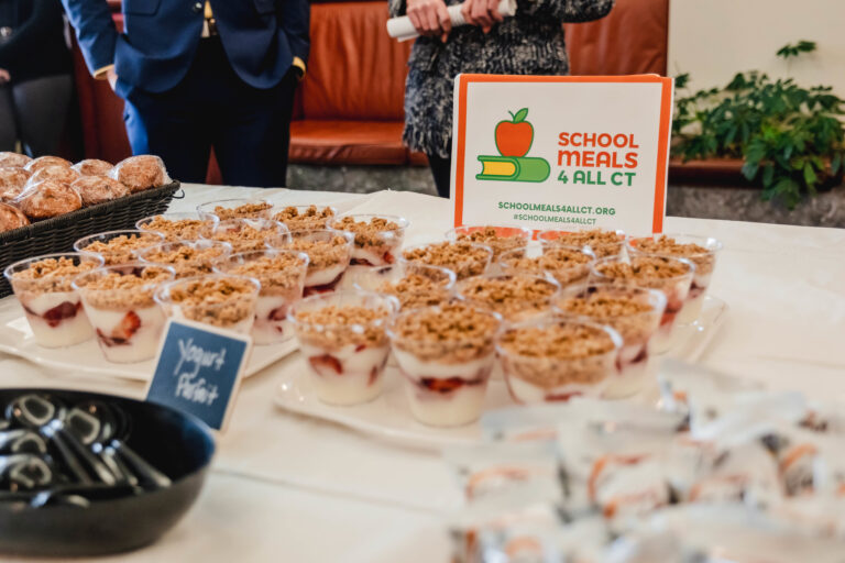 Dozens of small yogurt parfaits in clear plastic cups sit on trays on a table. Behind them on the table is a white and orange sign reading “SCHOOL MEALS 4 ALL CT,” and people dressed in business attire before a policy meeting are vaguely visible in the background.