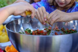 Children's hands squeeze citrus into a large bowl of garden-fresh salsa at a parent-led garden supper event.
