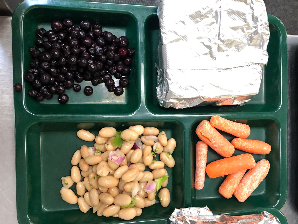 A green lunch tray with a foil-wrapped sandwich, a portion of blueberries, a portion of bean salad and a portion of baby carrots. 