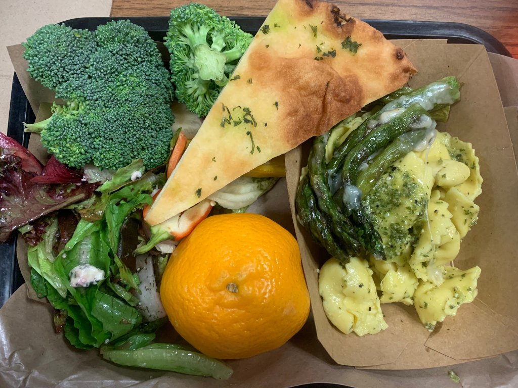 A black school lunch tray featuring a small dish of pesto pasta and asparagus, an orange, salad greens, and a piece of bread. 