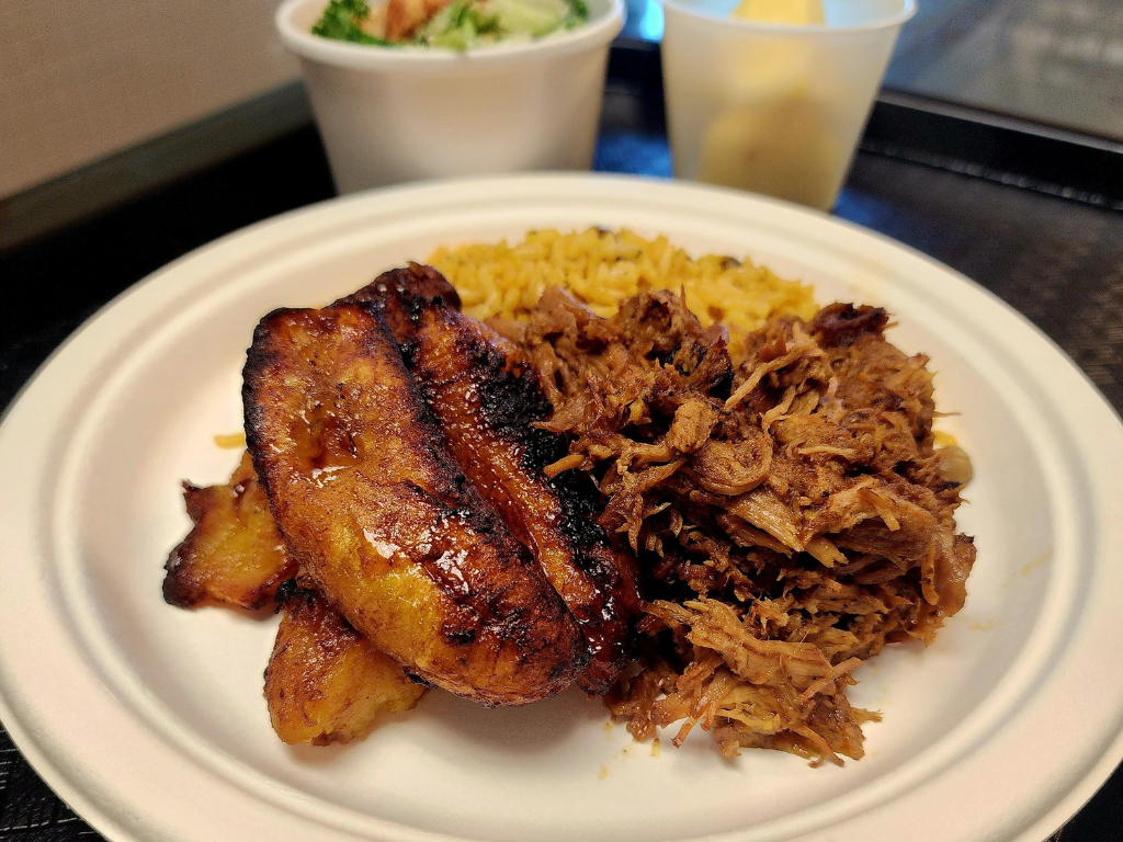A white plate with a serving of pork, rice and plantains, with a small salad in the background. 