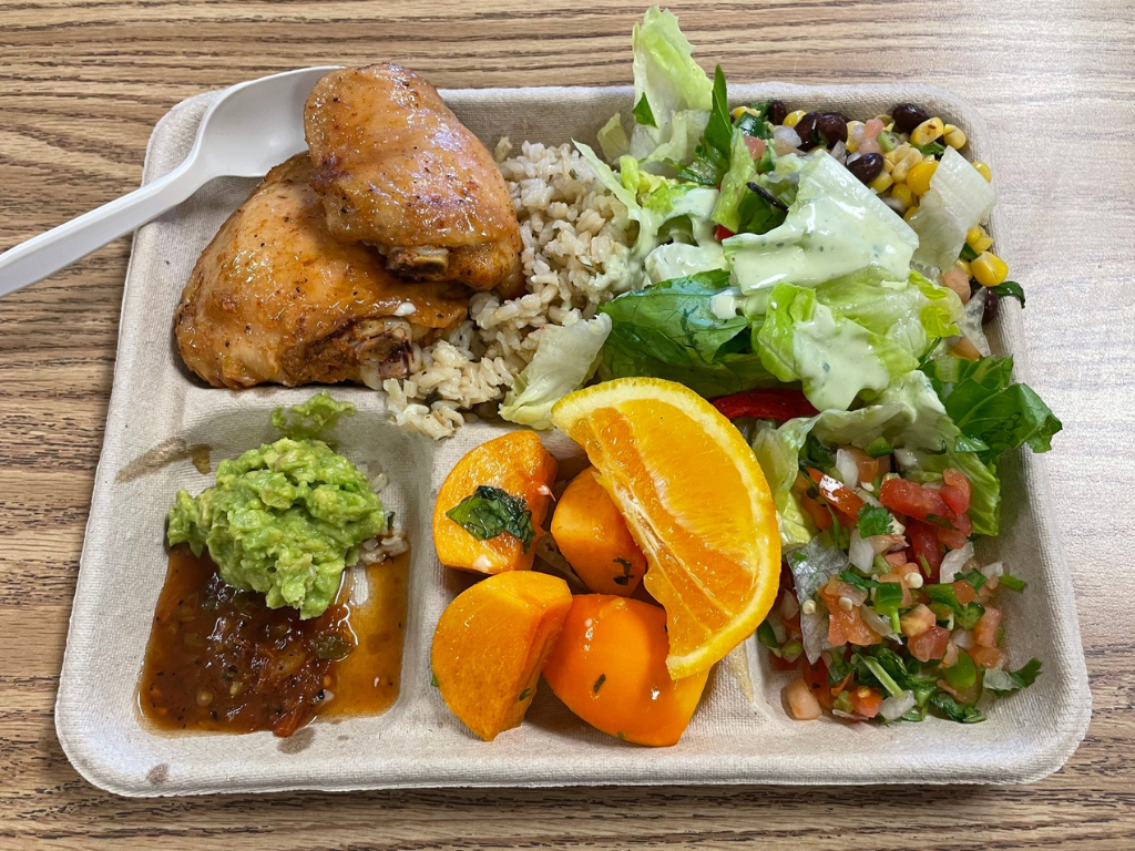 A beige school lunch tray on a wooden lunch table. On the tray are a chicken thigh sitting atop brown rice, orange and persimmon, a dollop of guacamole and sauce, and an array of local veggies. 