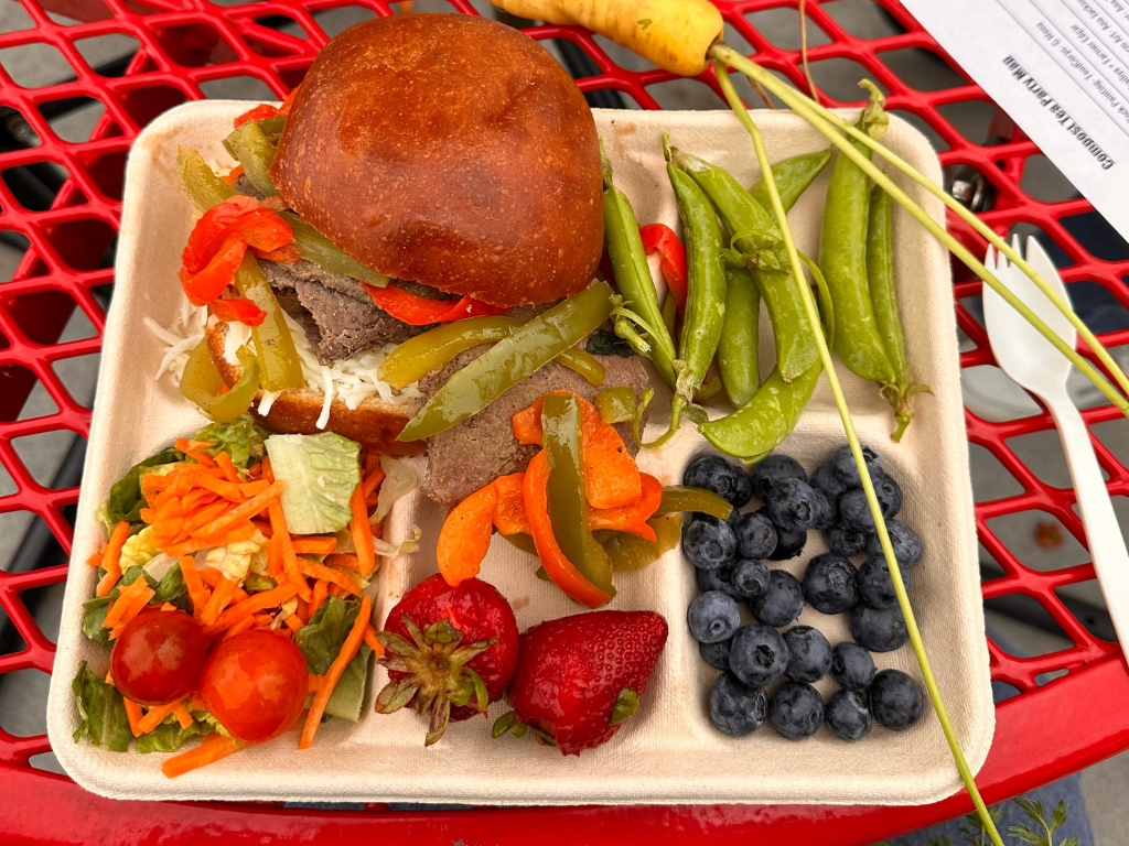 A beige school lunch tray placed on a red outdoor table. On the tray is a fajita steak and pepper sandwich, a handful of snap peas, a small salad, and some locally sourced strawberries and carrots. 
