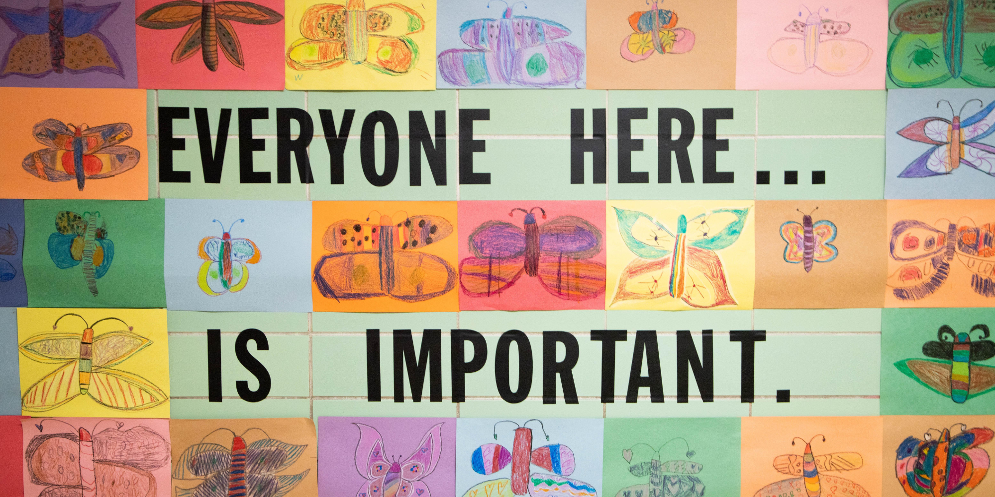 On a green school wall, large black text reads “EVERYONE HERE IS IMPORTANT.” The words are surrounded by colorful pictures of butterflies drawn by students. Messages like these are important for students to hear during LGBTQ Pride Month and all year long.