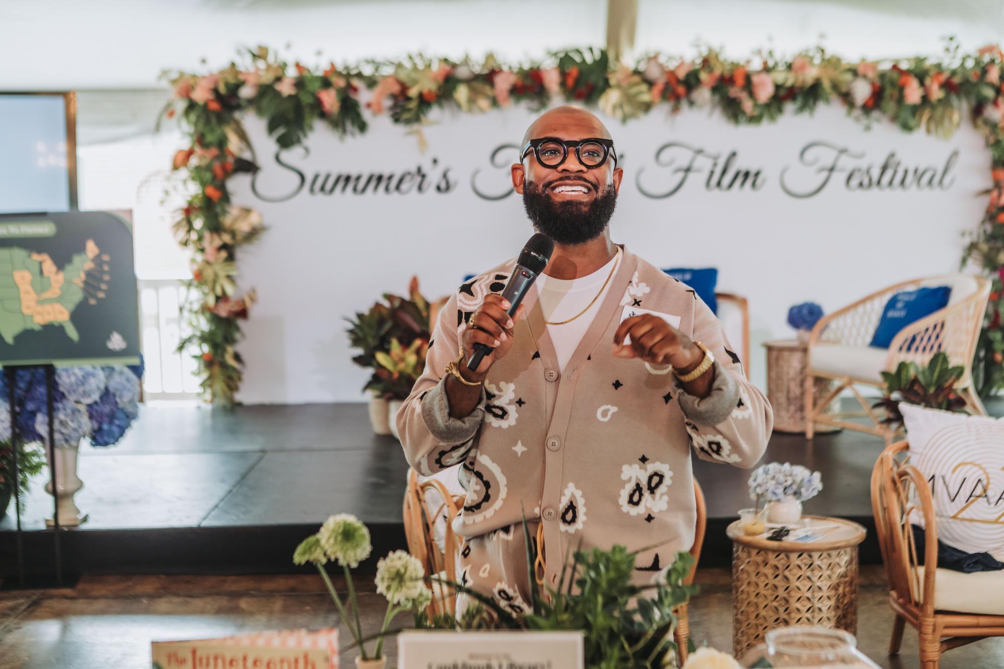 Dr. Robert S. Harvey stands in front of Martha’s Vineyard signs and behind a table of florals and other decor. He is holding a microphone and smiling as he speaks to a crowd at the Martha’s Vineyard African American Film Festival as part of FoodCorps’ Summer Movement Building Series.