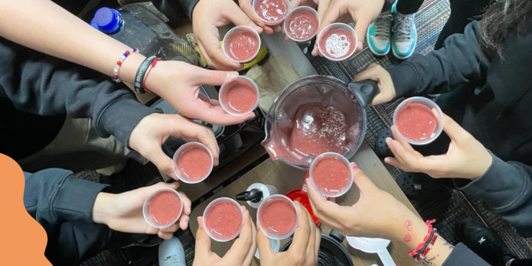 A dozen hands hold small sample-size cups of bright red smoothie in a circle. Below them is a blender and a series of scattered ingredients.