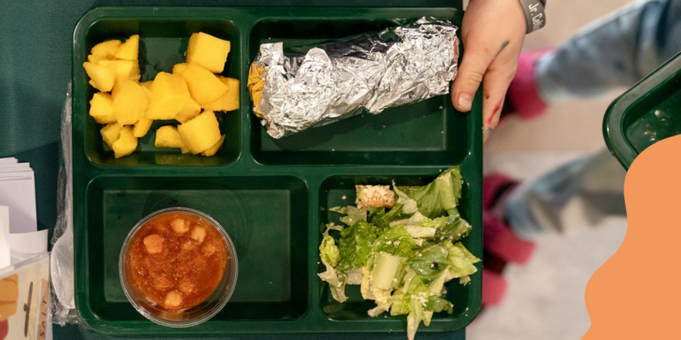 A student’s hands holding a green lunch tray, carrying a wrap in foil, cubed mango, a side salad and a taste-test sized cup of stew. CEP allows more students to access school meals for free.