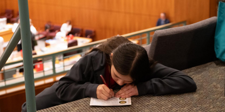 A student wearing a dark hoodie sits on a carpeted step in New Mexico’s Roundhouse and writes on a postcard about school food policy. Behind them, the legislature is in session.