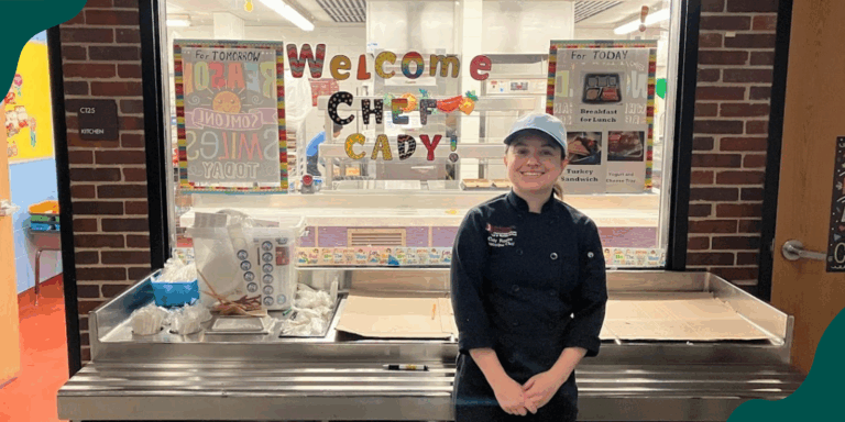 FoodCorps alumni Cady Molloy wears a black chef's coat and baseball cap, standing in front of a school kitchen with "Welcome Chef Cady!" printed in colorful letters on the glass.
