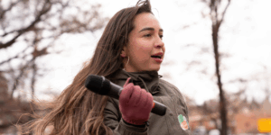 Taty Trujillo wears an olive-green coat, red gloves, and gold hoop earrings, and has long brown hair blowing behind her. She is looking off-camera, holding a microphone, and addressing an outdoor crowd at the New Mexico state legislature.