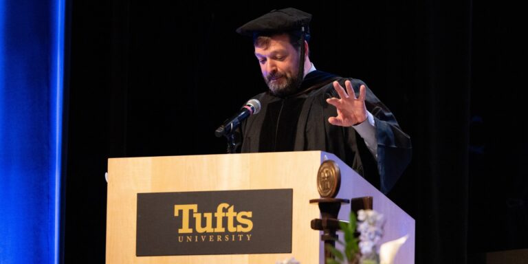 Curt Ellis in academic regalia delivers a commencement address from a podium featuring the Tufts University plaque.