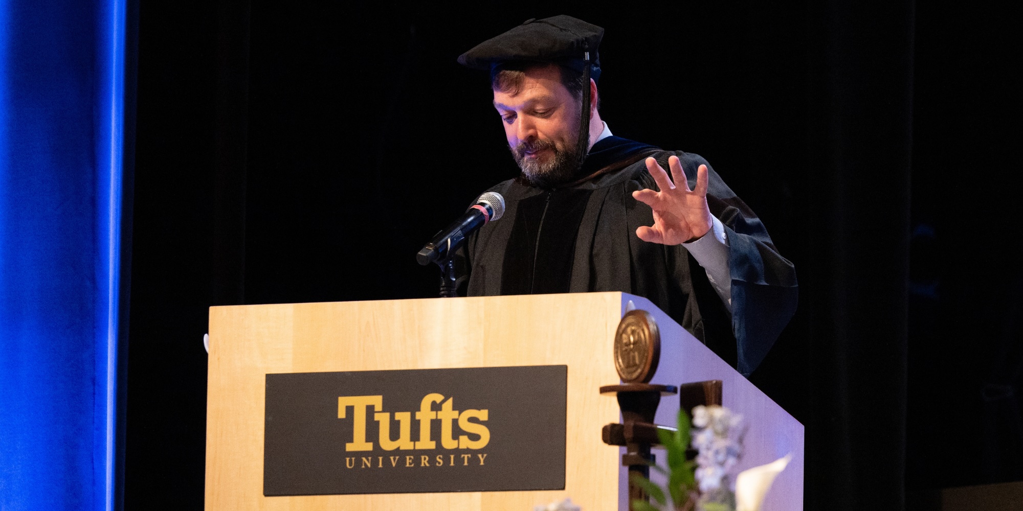 Curt Ellis in academic regalia delivers a commencement address from a podium featuring the Tufts University plaque.